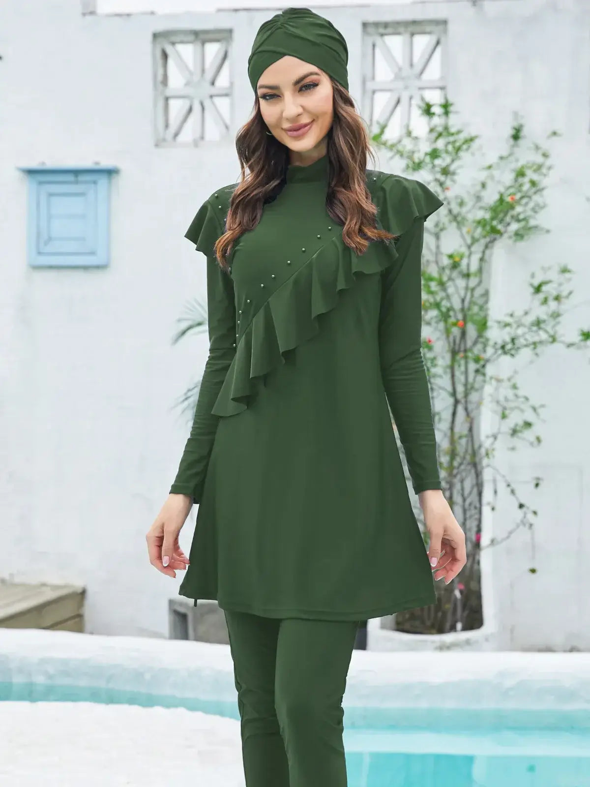 Woman wearing green modest burkini with hijab, long-sleeve top, and pants, standing by pool. Muslim swimwear for women.