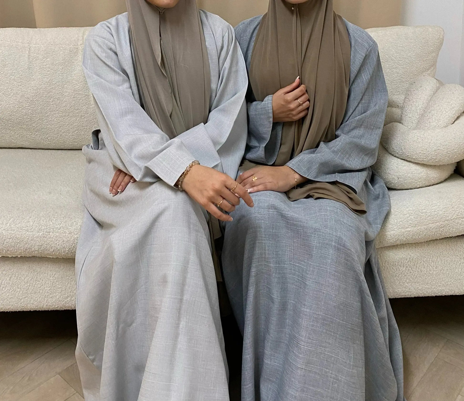 Two women wearing luxurious closed linen abayas in a classic silhouette, showcasing Dubai's timeless fashion, seated on a white sofa.