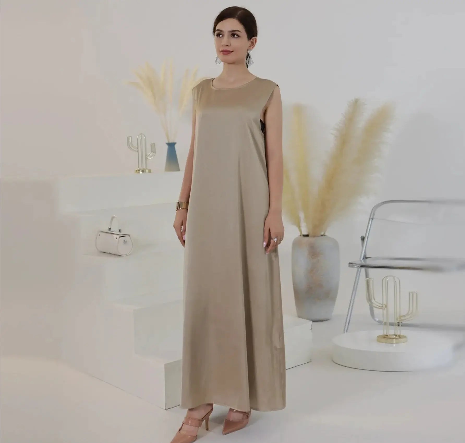 Woman wearing beige sleeveless inner abaya dress standing in a minimalist room with modern decor.