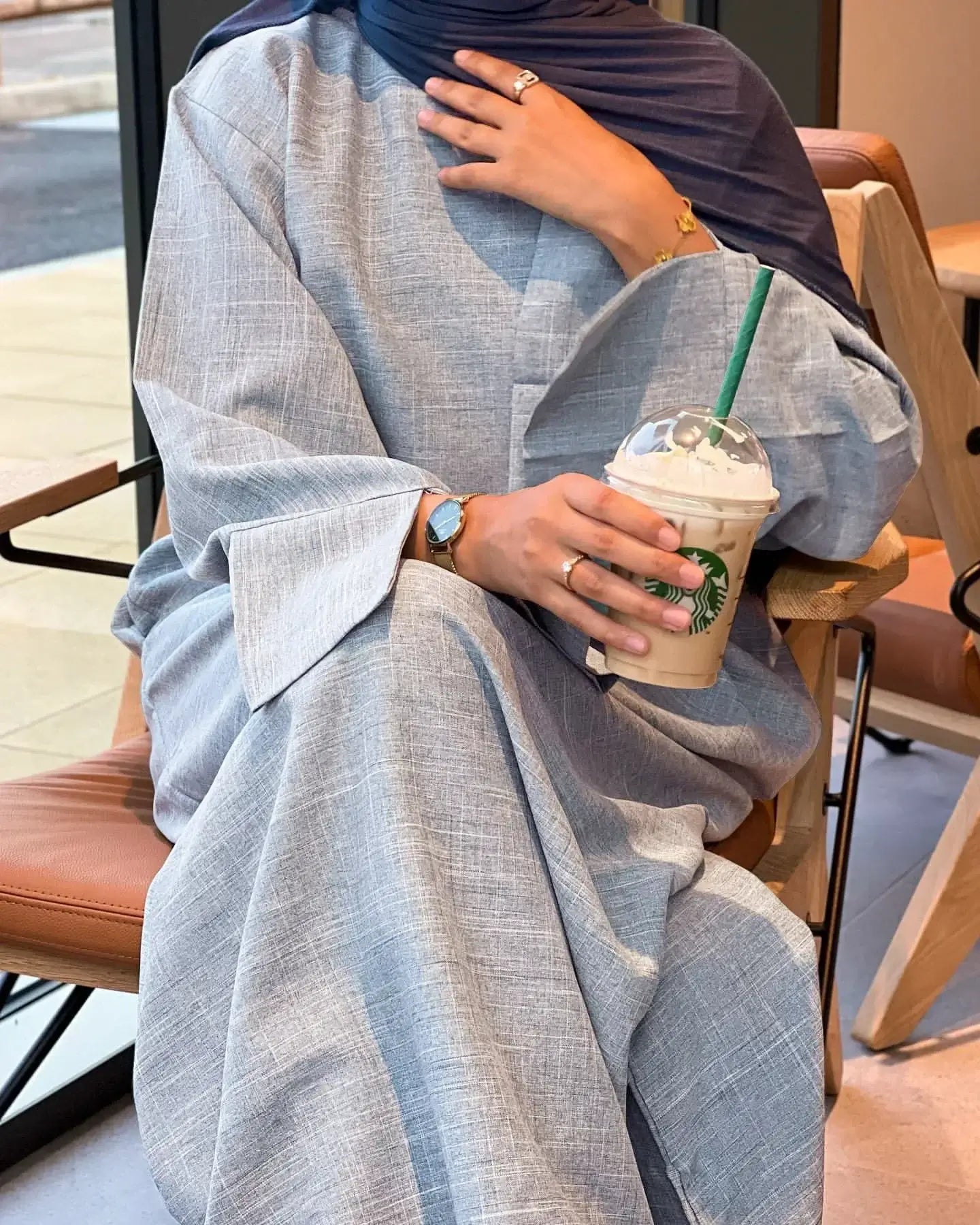 Woman wearing a luxurious closed linen abaya, holding a Starbucks iced coffee in a modern cafe.