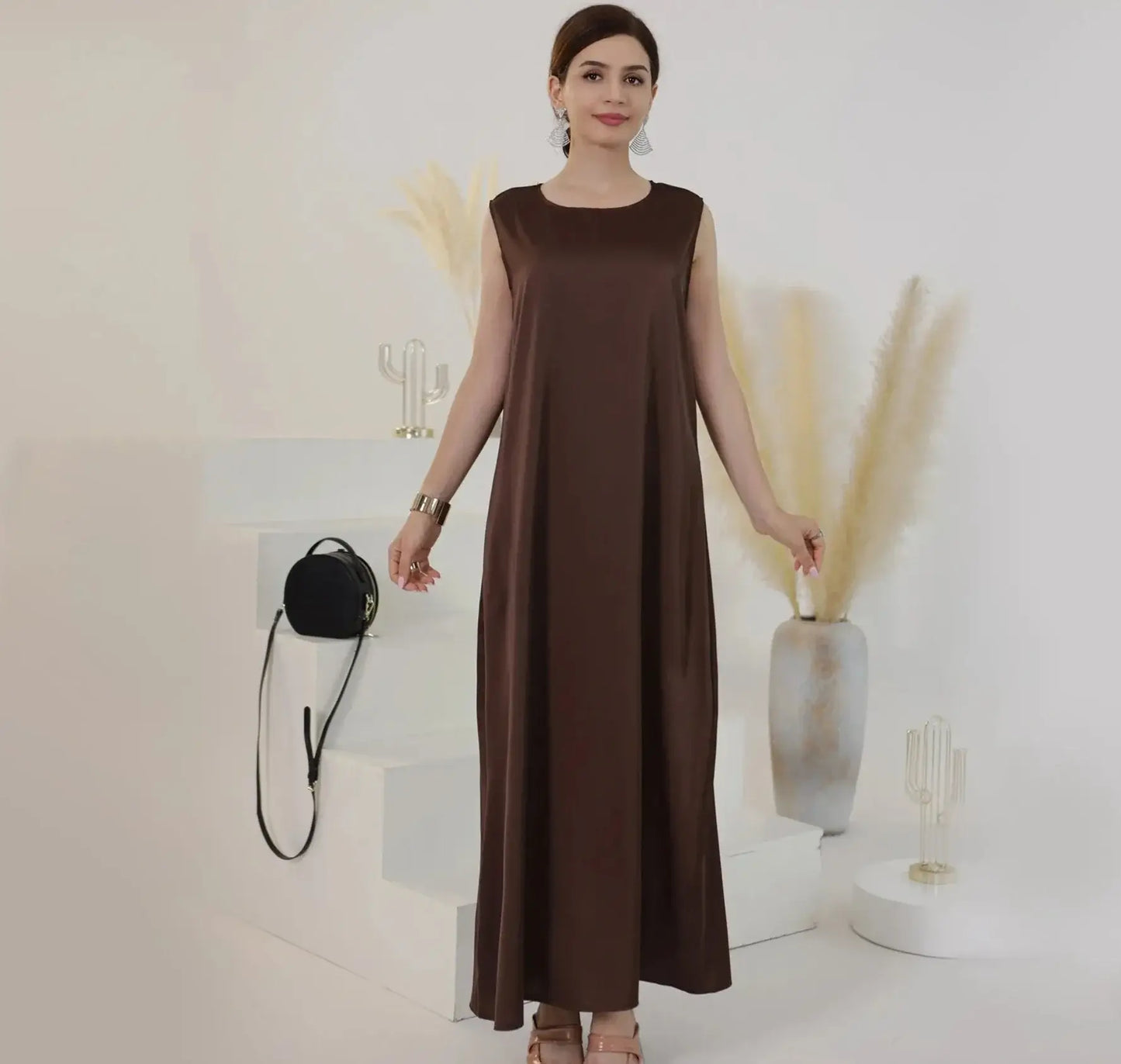 Woman wearing a brown sleeveless long dress, standing against a minimalist backdrop with neutral decor.