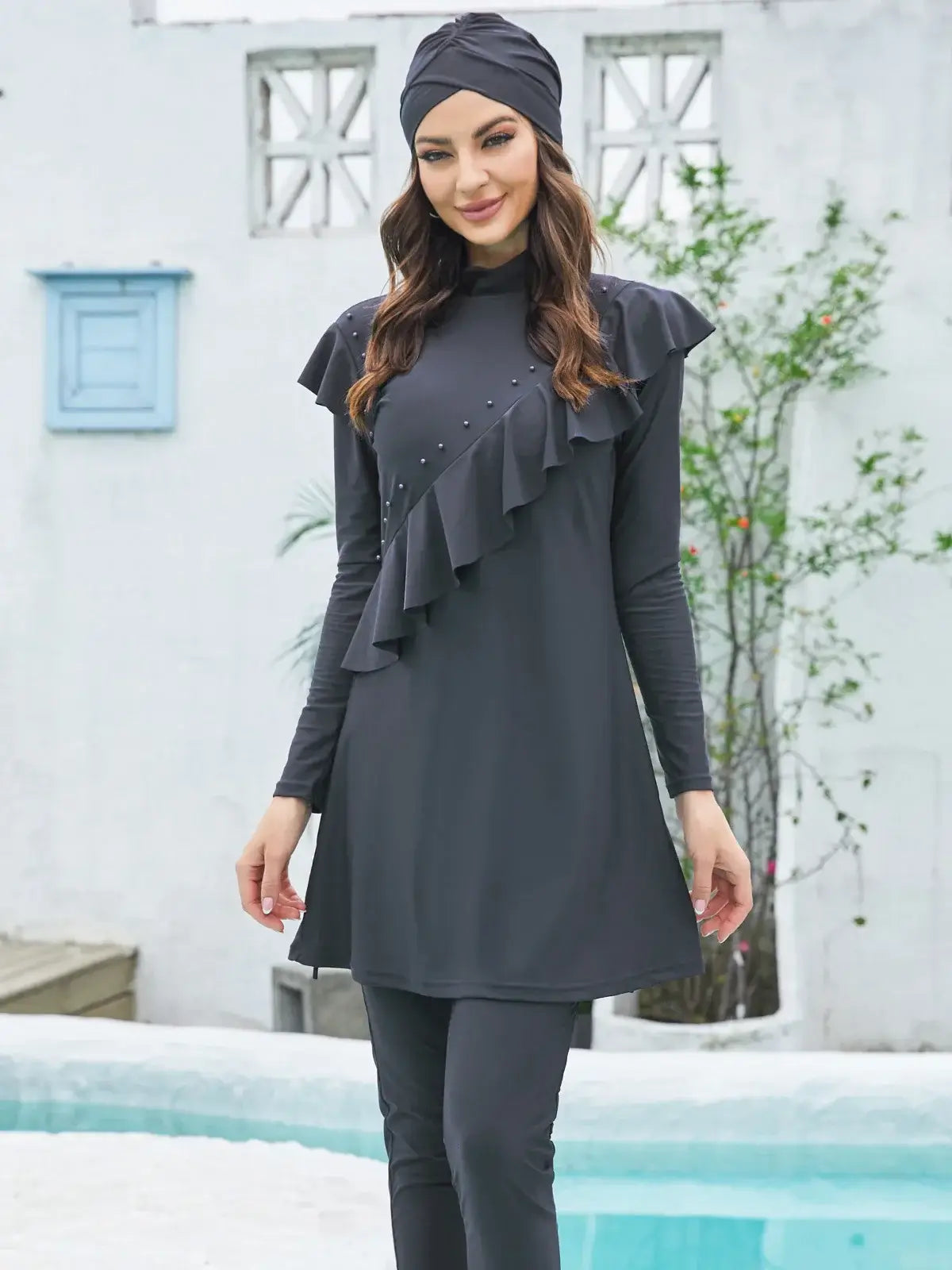 Woman wearing stylish modest black burkini set with long-sleeve top, pants, and hijab near a pool. Premium spandex and polyester swimwear.