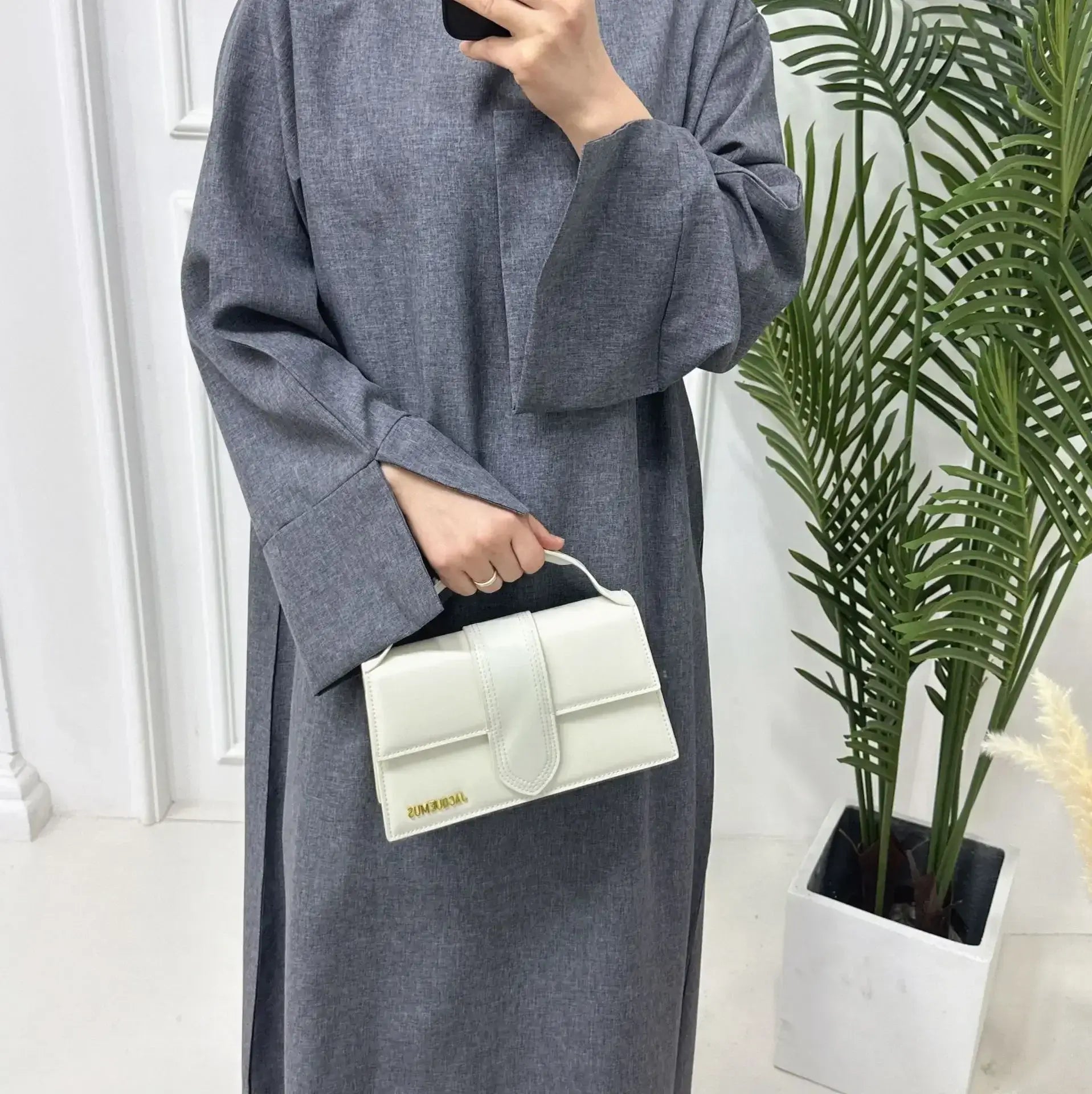 Woman wearing grey closed linen abaya with white handbag, standing in elegant indoor setting with plants.