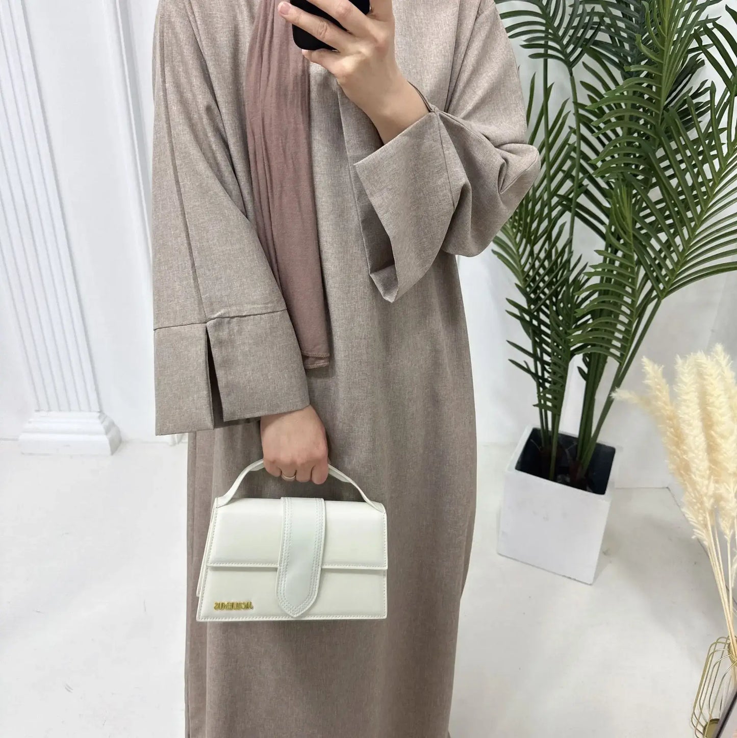 Woman wearing a closed linen abaya holding a white handbag in a modern, minimalist setting