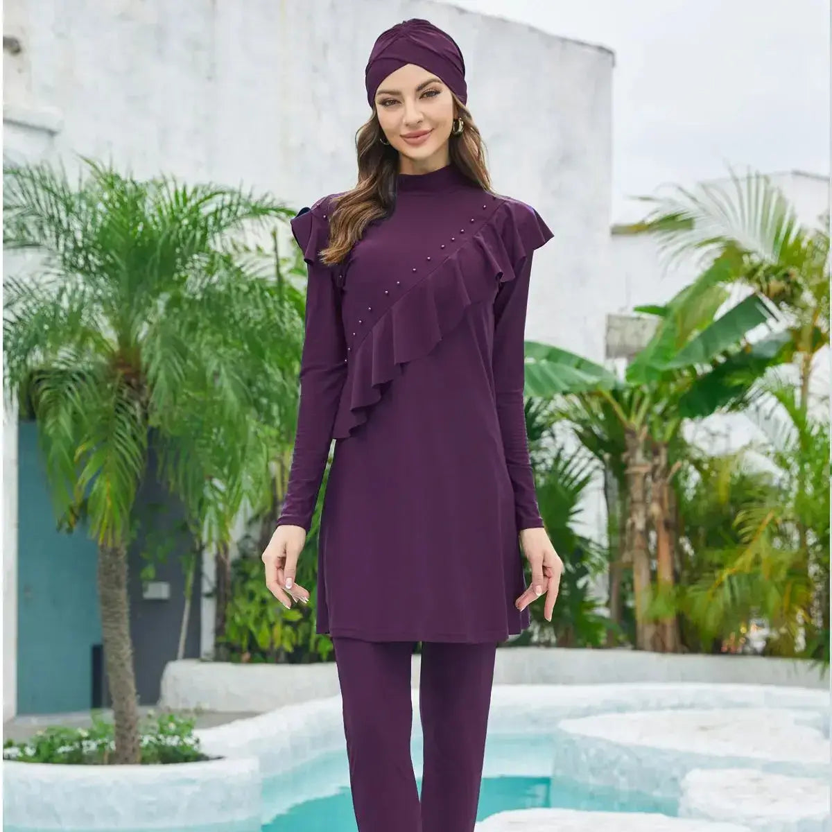 Woman wearing purple Lake Modest Burkini with hijab, top, and pants, standing by a pool with palm trees in the background.