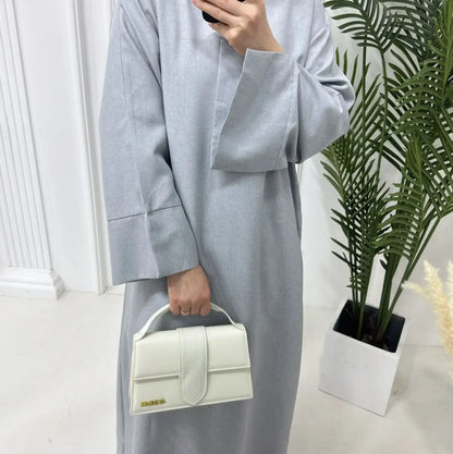 Woman wearing a grey closed linen abaya holding a white handbag, standing indoors next to green plants.