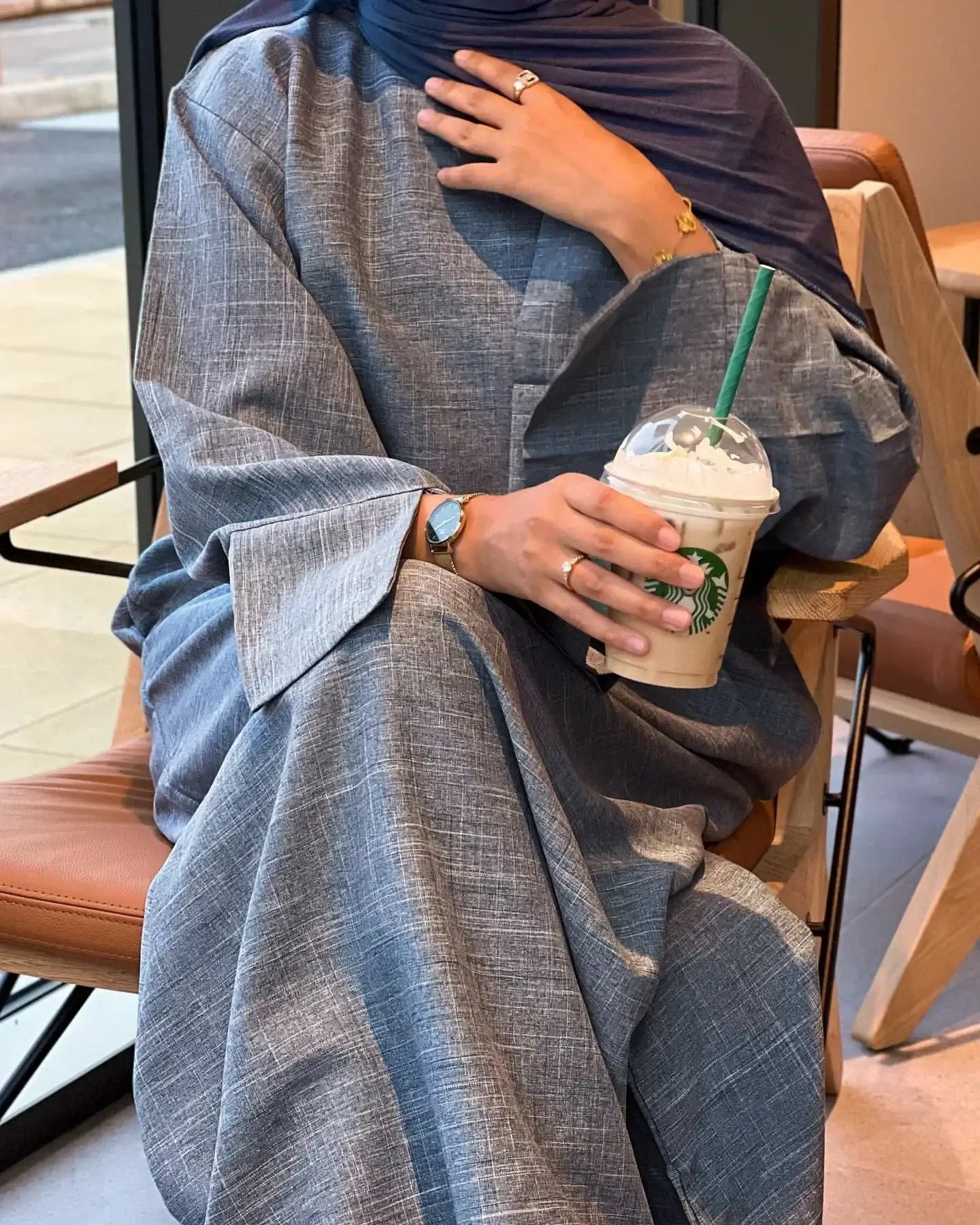 Woman wearing Closed Linen Abaya enjoying a Starbucks drink indoors, showcasing the luxurious fabric and sophisticated design.